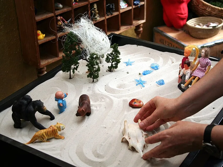 Hands arranging miniature figures, trees and shells in a sand tray